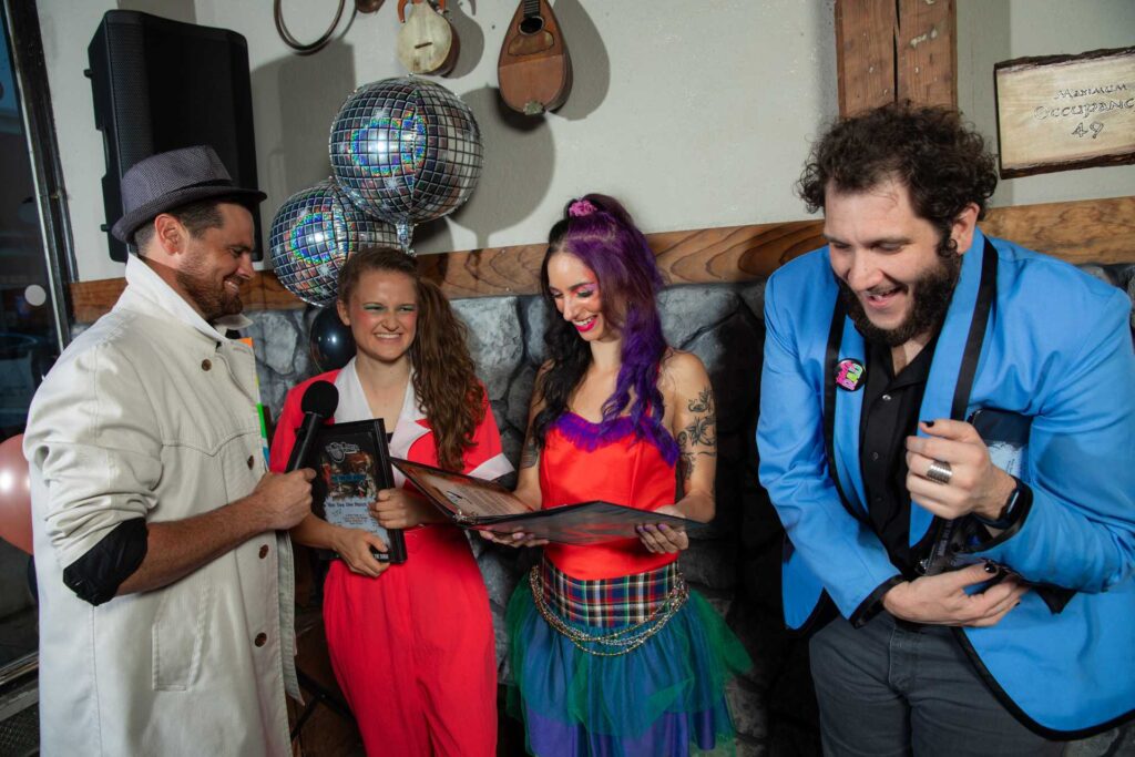 Four friends dressed in 1980s attire laugh while attending a 1980s-themed murder mystery party